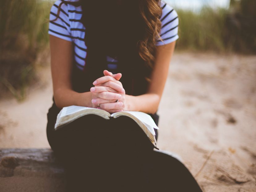 young girl in striy top with her hands clasped praying with a bible on her knee