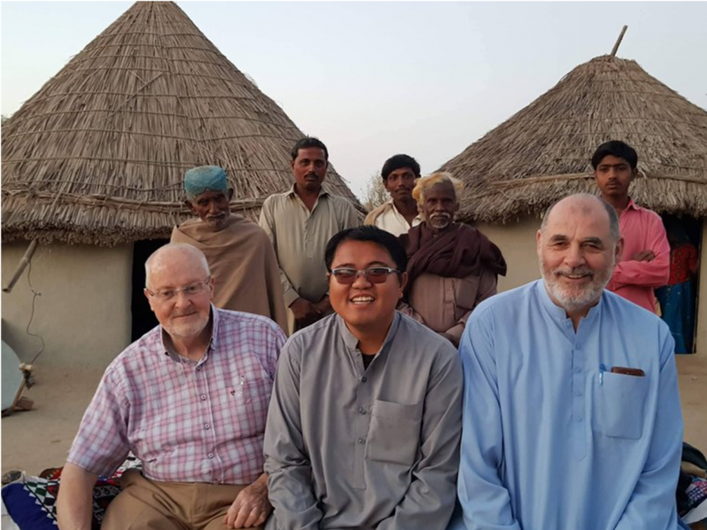 Men smiling at the camera in front of Pakistani homes